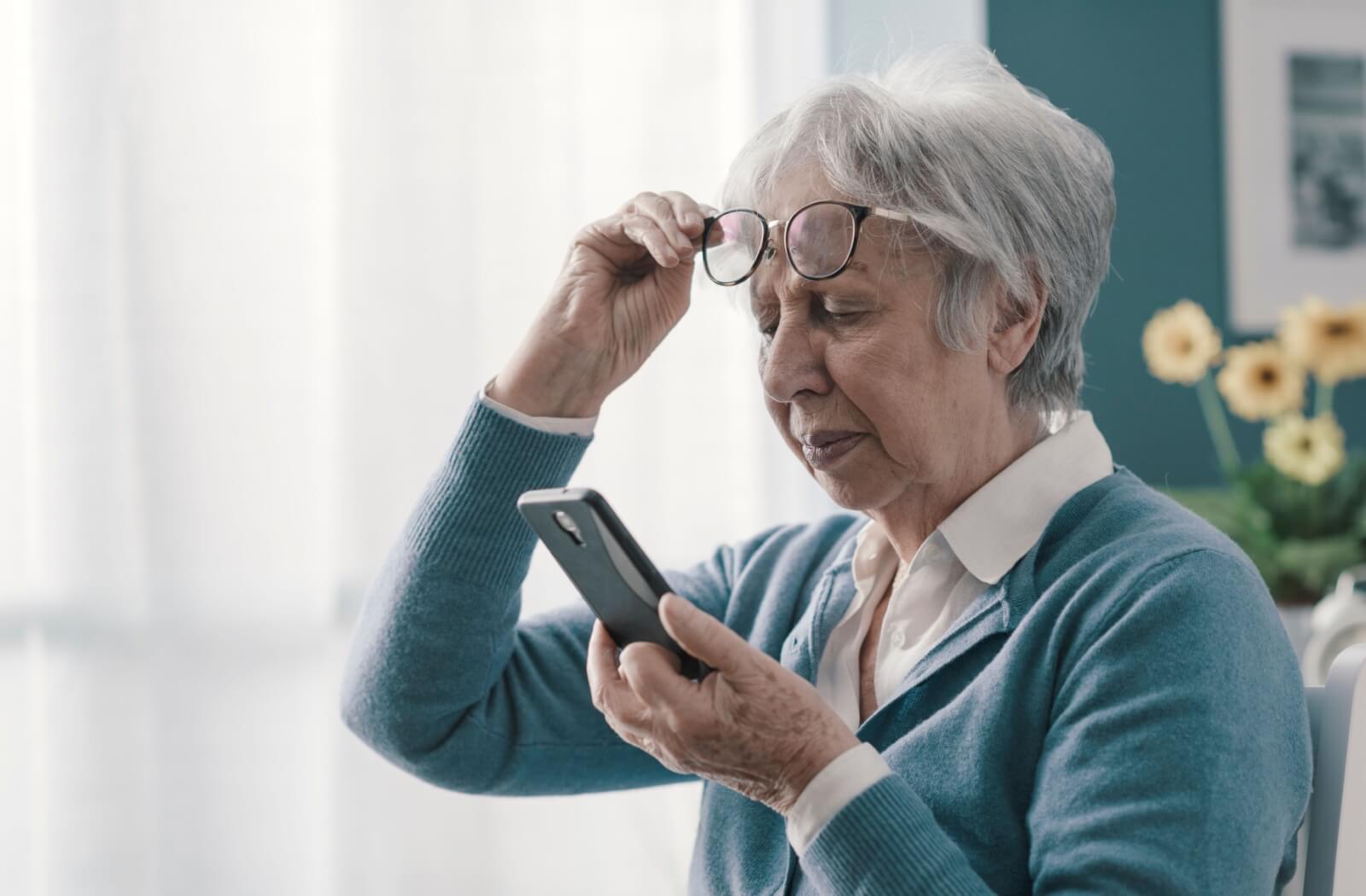 Older woman adjusting glasses while looking at her smartphone indoors.