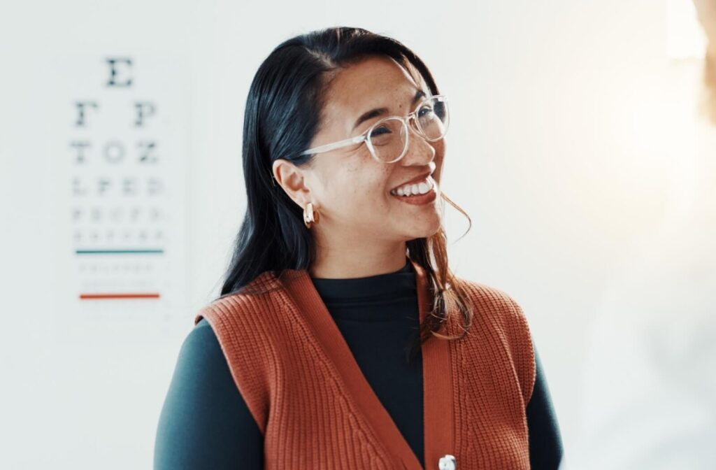 A person with dark hair wearing clear-rimmed eyeglasses and a burnt orange vest smiling during an eye exam, illustrating the benefits of OptiLight for managing dry eye symptoms.