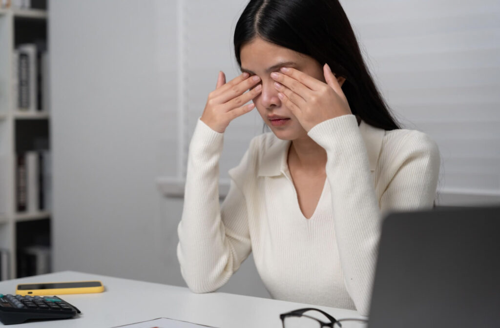 A person sitting at a white desk rubbing their closed eyes with both hands, illustrating symptoms of morning dry eye and digital eye strain.